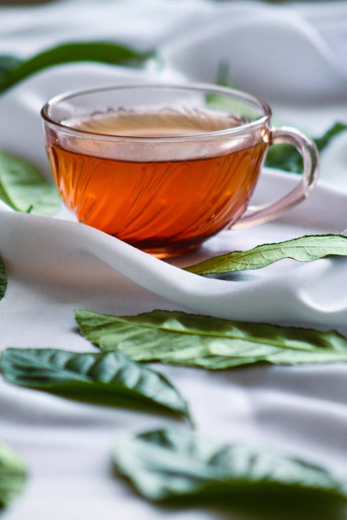 Close-up of tea in a glass cup surrounded by green leaves on a white cloth.